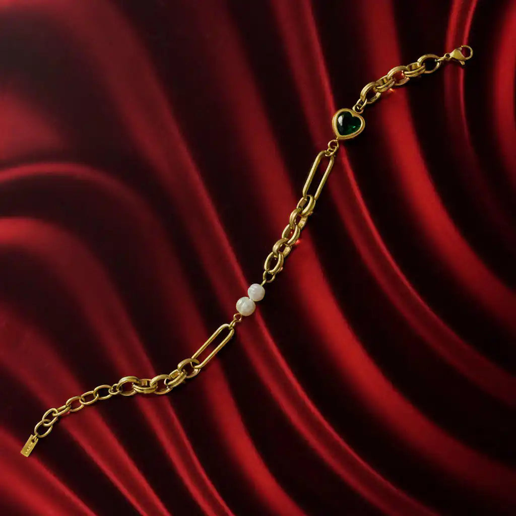 Gold-toned chain bracelet featuring a vibrant green heart-shaped gem, two lustrous white pearls, and a mix of oval and paperclip-style links displayed on a flowing rich red velvet fabric background