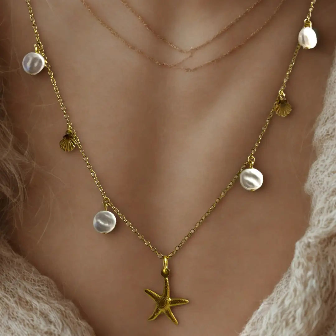 A close-up of a gold necklace featuring irregular white pearls, tiny gold scallop shells, and a central starfish pendant, worn by a person over a soft cream knit top