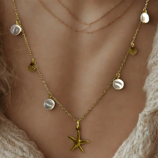 A close-up of a gold necklace featuring irregular white pearls, tiny gold scallop shells, and a central starfish pendant, worn by a person over a soft cream knit top